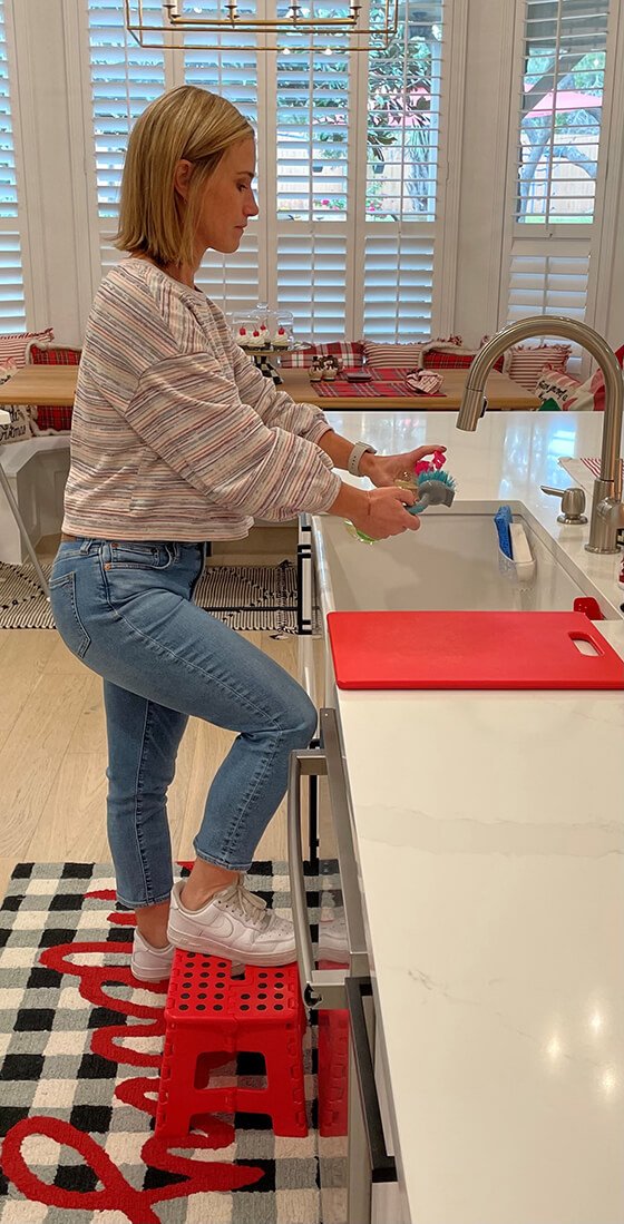 Woman standing at the kitchen sink, with one foot on a step stool.