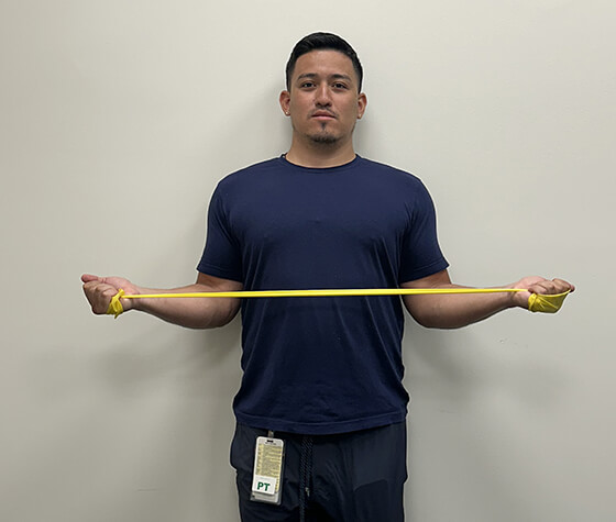 A male physical therapist stretching an exercise band outwardly past his shoulders. 