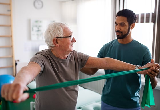 An elderly man using an exercise band for his back muscles with assistance from a physical therapist. 